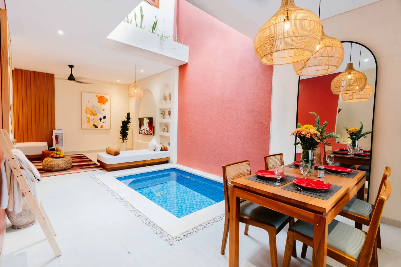 Compact white-tiled bathroom with round black mirror, woven pendant light, frangipani flowers on shelf, white sink, toilet, and folded towels. - Bali Villas