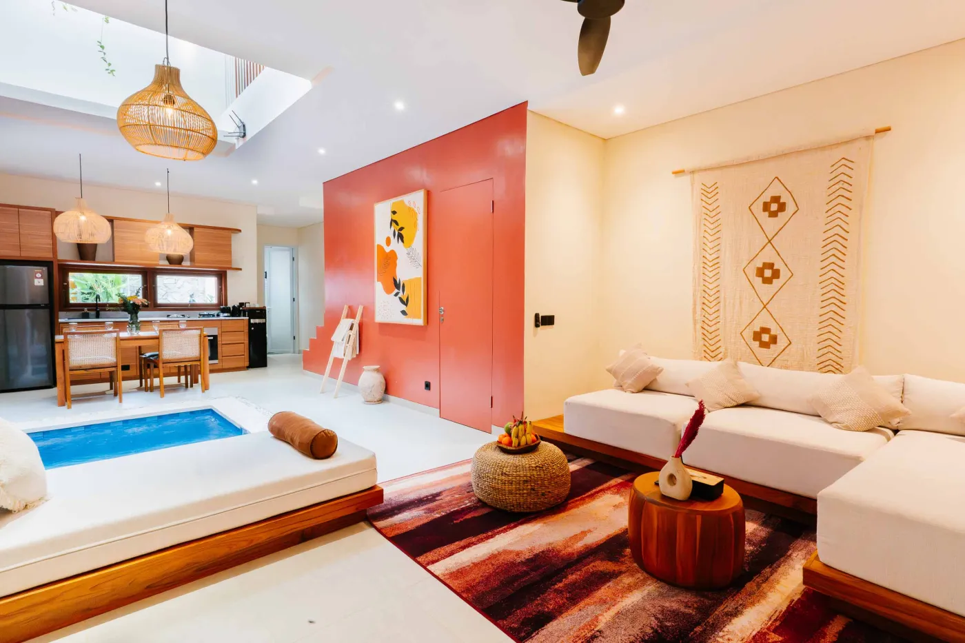 Compact white-tiled bathroom with round black mirror, woven pendant light, frangipani flowers on shelf, white sink, toilet, and folded towels. - Bali Villas