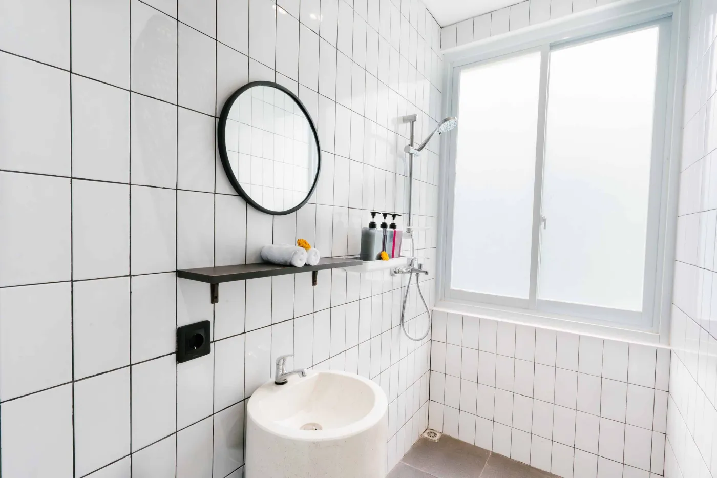 Compact white-tiled bathroom with round black mirror, woven pendant light, frangipani flowers on shelf, white sink, toilet, and folded towels. - Bali Villas