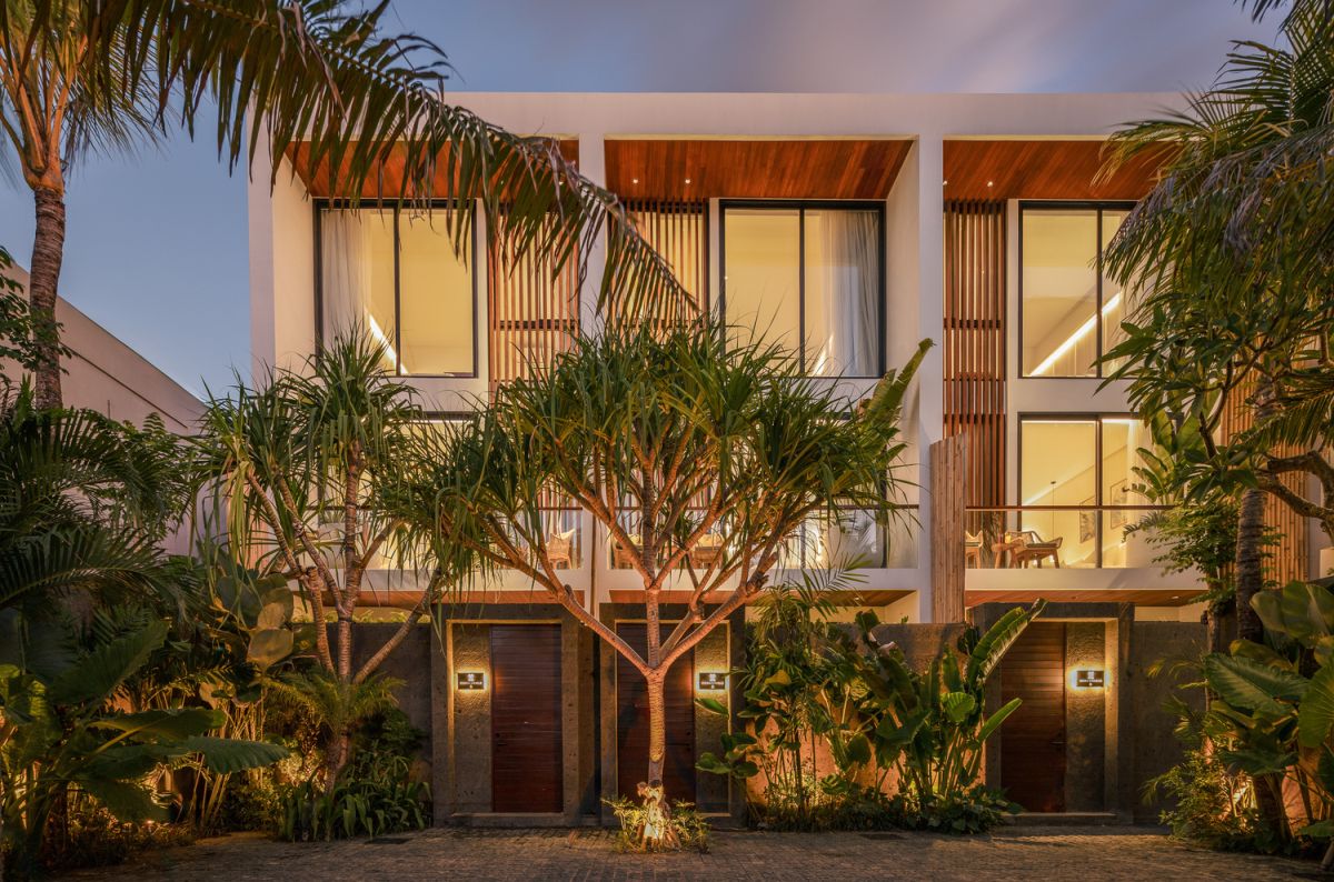 Modern open-plan kitchen and living room interior with vertical green wall, wooden staircase, bar stools, and plush green sofa against white marble flooring. - Bali Villas