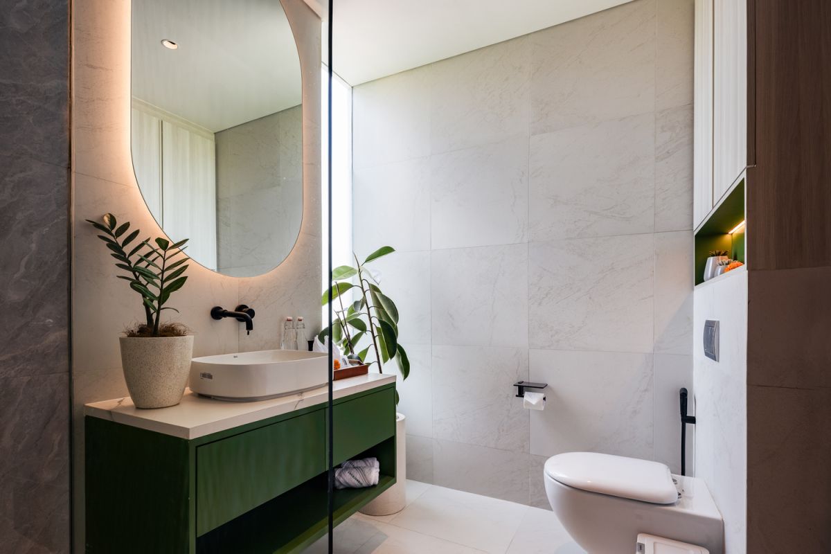 Modern open-plan kitchen and living room interior with vertical green wall, wooden staircase, bar stools, and plush green sofa against white marble flooring. - Bali Villas