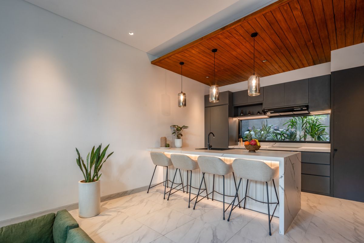 Modern open-plan kitchen and living room interior with vertical green wall, wooden staircase, bar stools, and plush green sofa against white marble flooring. - Bali Villas