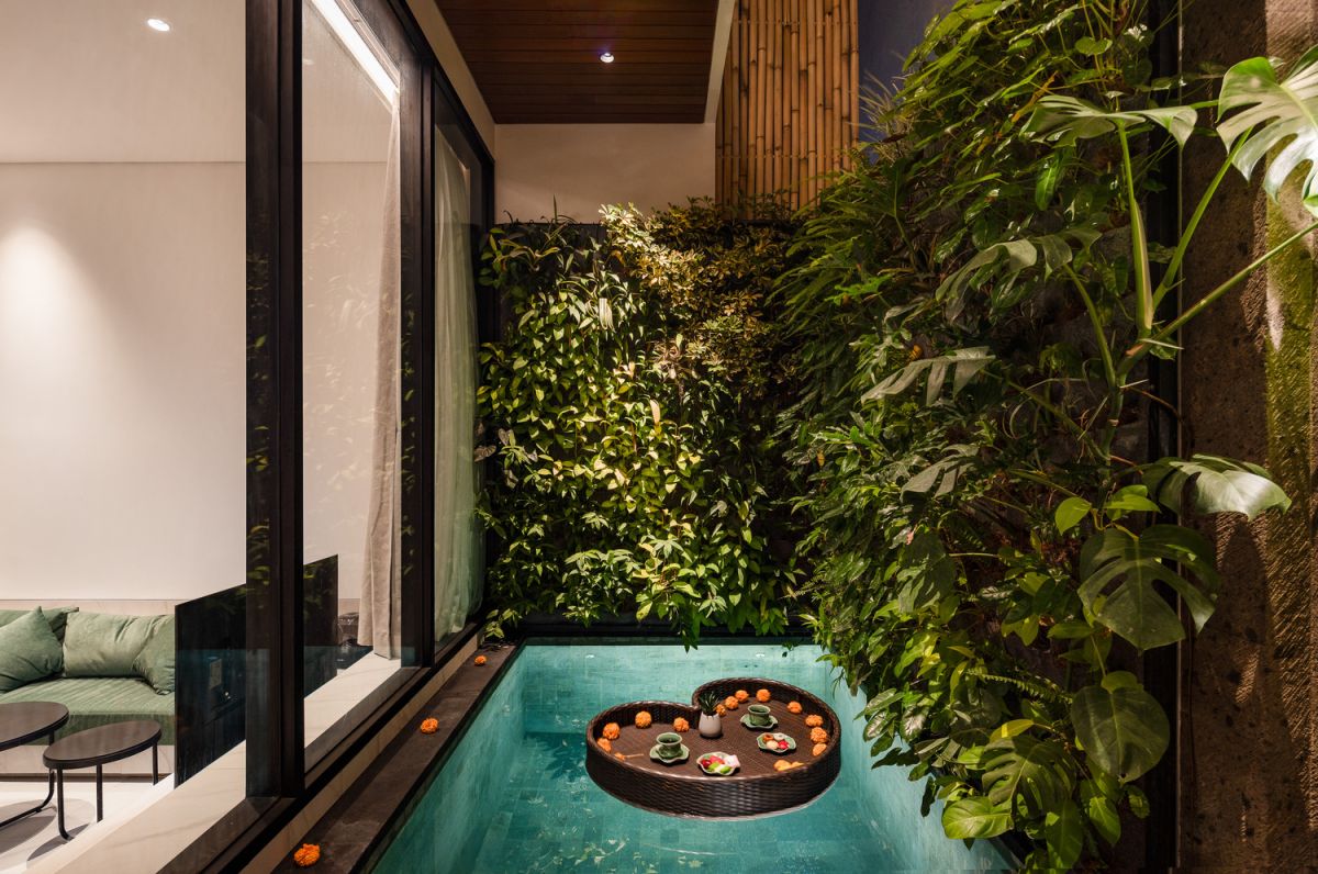 Modern open-plan kitchen and living room interior with vertical green wall, wooden staircase, bar stools, and plush green sofa against white marble flooring. - Bali Villas