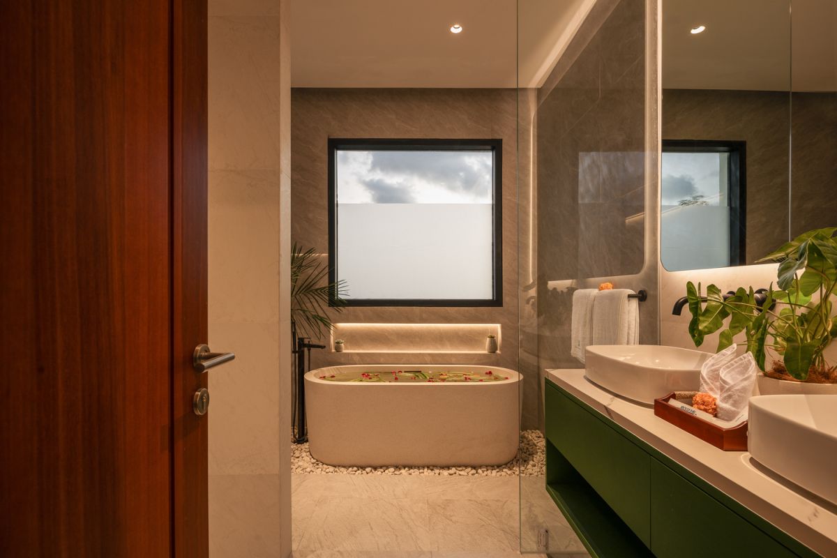 Modern open-plan kitchen and living room interior with vertical green wall, wooden staircase, bar stools, and plush green sofa against white marble flooring. - Bali Villas