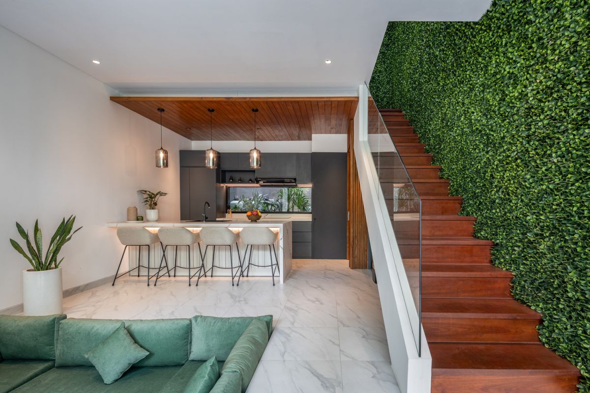 Modern open-plan kitchen and living room interior with vertical green wall, wooden staircase, bar stools, and plush green sofa against white marble flooring. - Bali Villas
