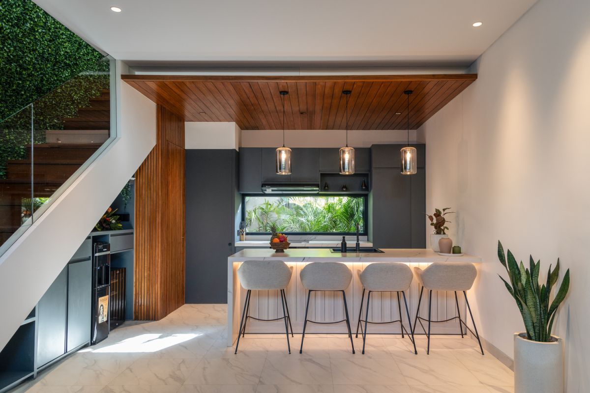 Modern open-plan kitchen and living room interior with vertical green wall, wooden staircase, bar stools, and plush green sofa against white marble flooring. - Bali Villas