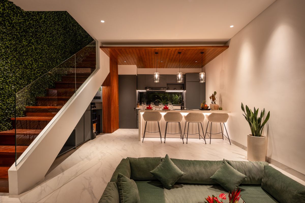 Modern open-plan kitchen and living room interior with vertical green wall, wooden staircase, bar stools, and plush green sofa against white marble flooring. - Bali Villas