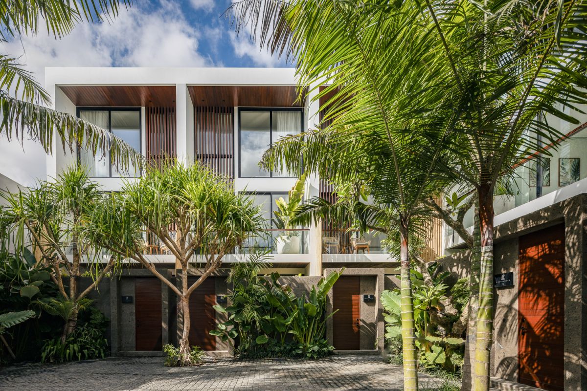 Modern open-plan kitchen and living room interior with vertical green wall, wooden staircase, bar stools, and plush green sofa against white marble flooring. - Bali Villas