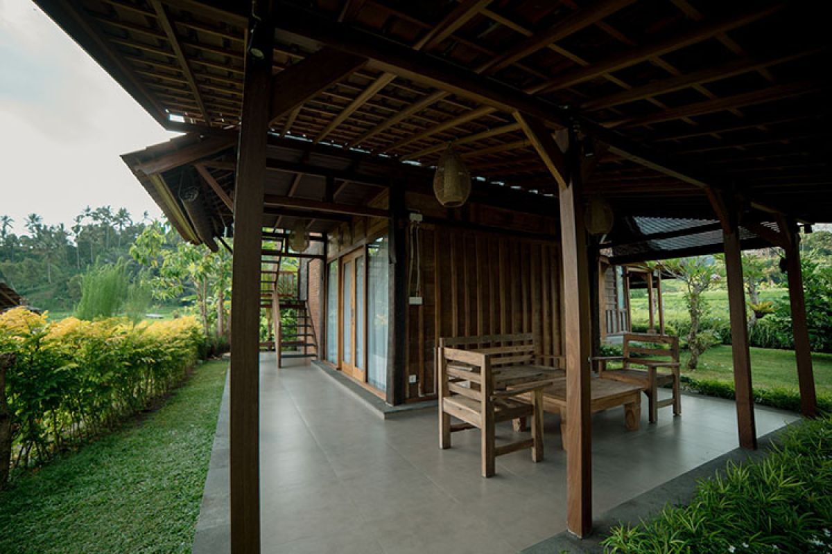 Traditional wooden Balinese bungalow with covered porch, benches, and table, surrounded by lush tropical gardens and rice paddies. - Bali Villas