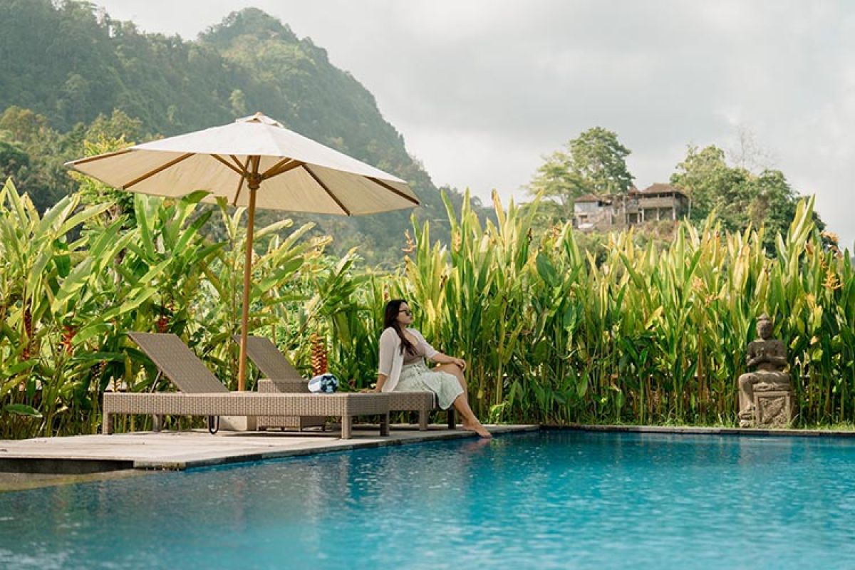 Traditional wooden Balinese bungalow with covered porch, benches, and table, surrounded by lush tropical gardens and rice paddies. - Bali Villas