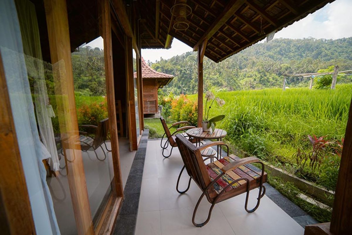 Traditional wooden Balinese bungalow with covered porch, benches, and table, surrounded by lush tropical gardens and rice paddies. - Bali Villas