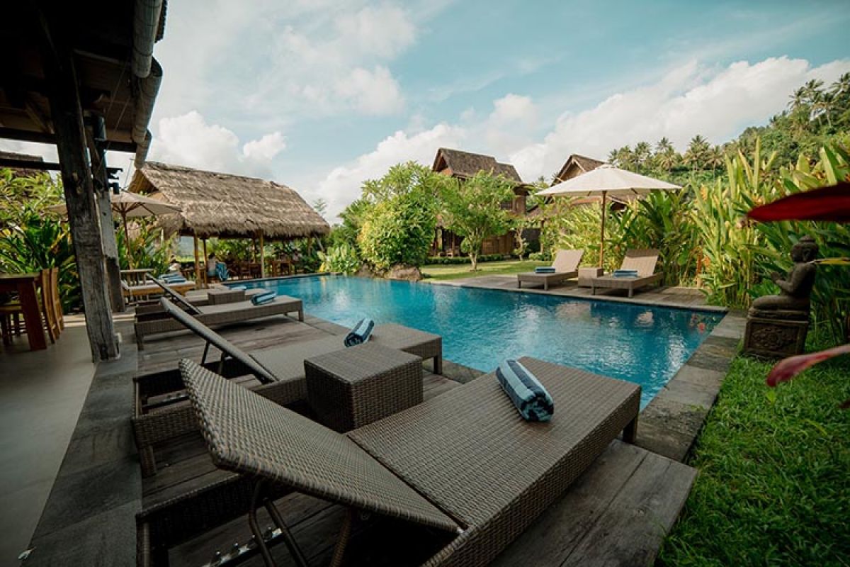 Traditional wooden Balinese bungalow with covered porch, benches, and table, surrounded by lush tropical gardens and rice paddies. - Bali Villas