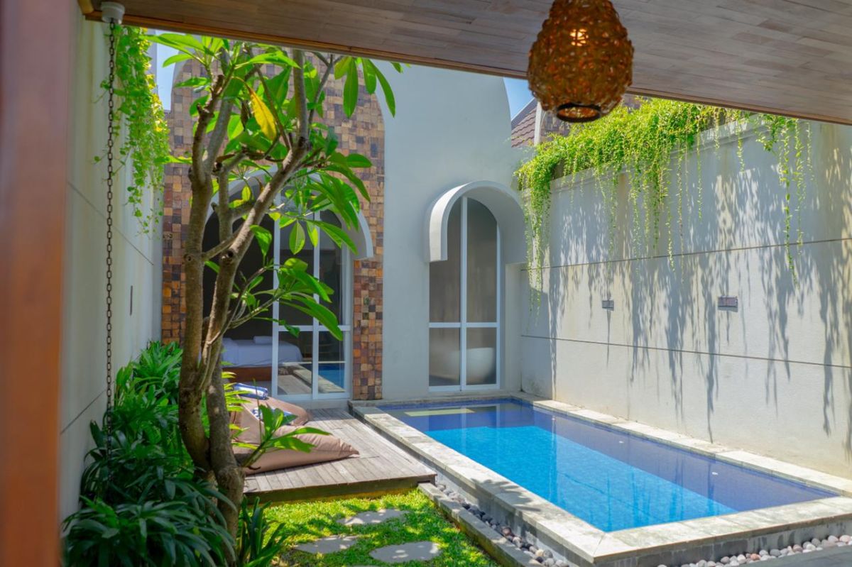 Modern interior with white bathtub and open archway leading to a private blue infinity pool, wooden lounge chairs, hanging lantern, and lush green vines in a serene courtyard. - Bali Villas