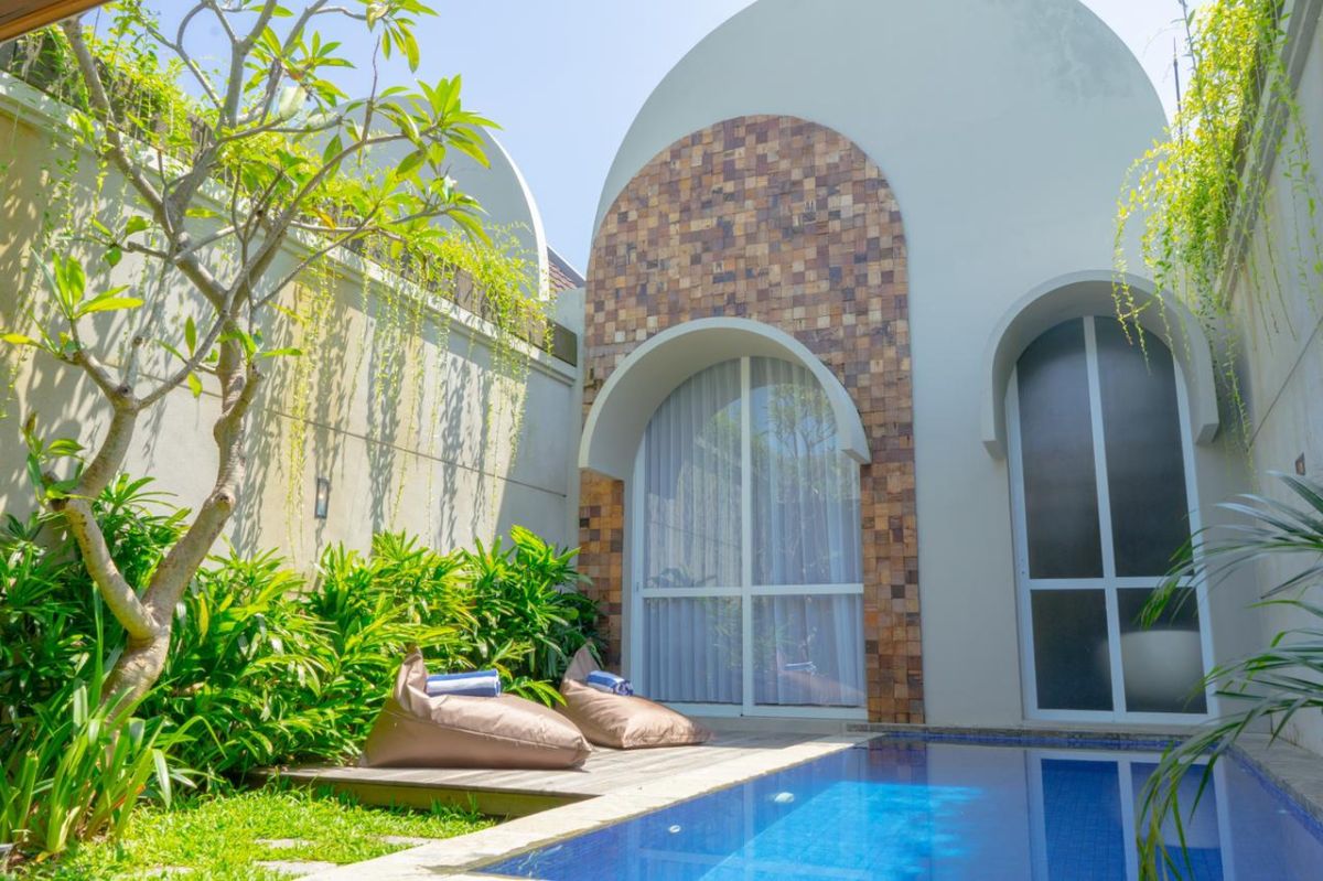 Modern interior with white bathtub and open archway leading to a private blue infinity pool, wooden lounge chairs, hanging lantern, and lush green vines in a serene courtyard. - Bali Villas