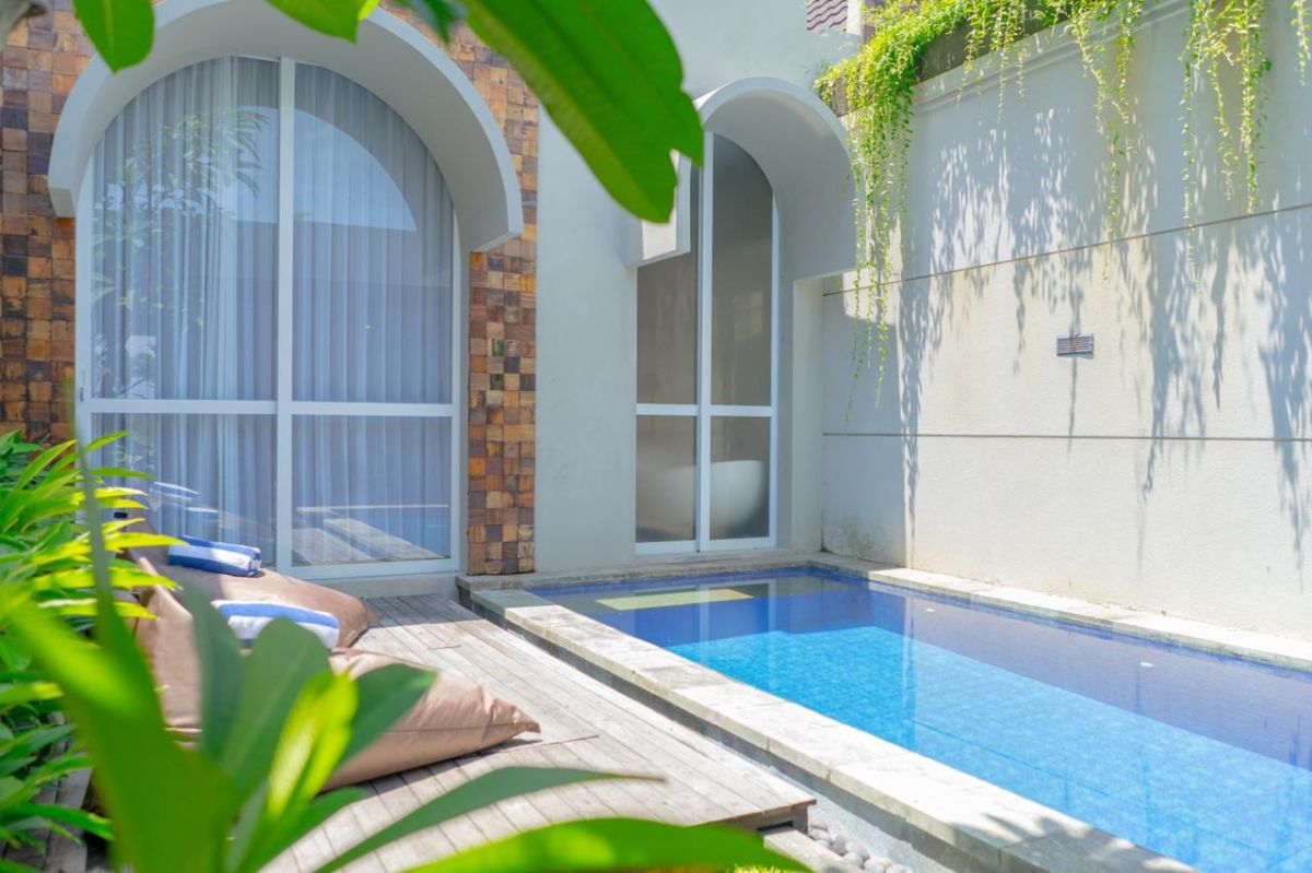 Serene tropical villa with private infinity-edge pool, lush surrounding greenery, arched windows, and cushioned lounge chairs on a wooden deck. - Bali Villas