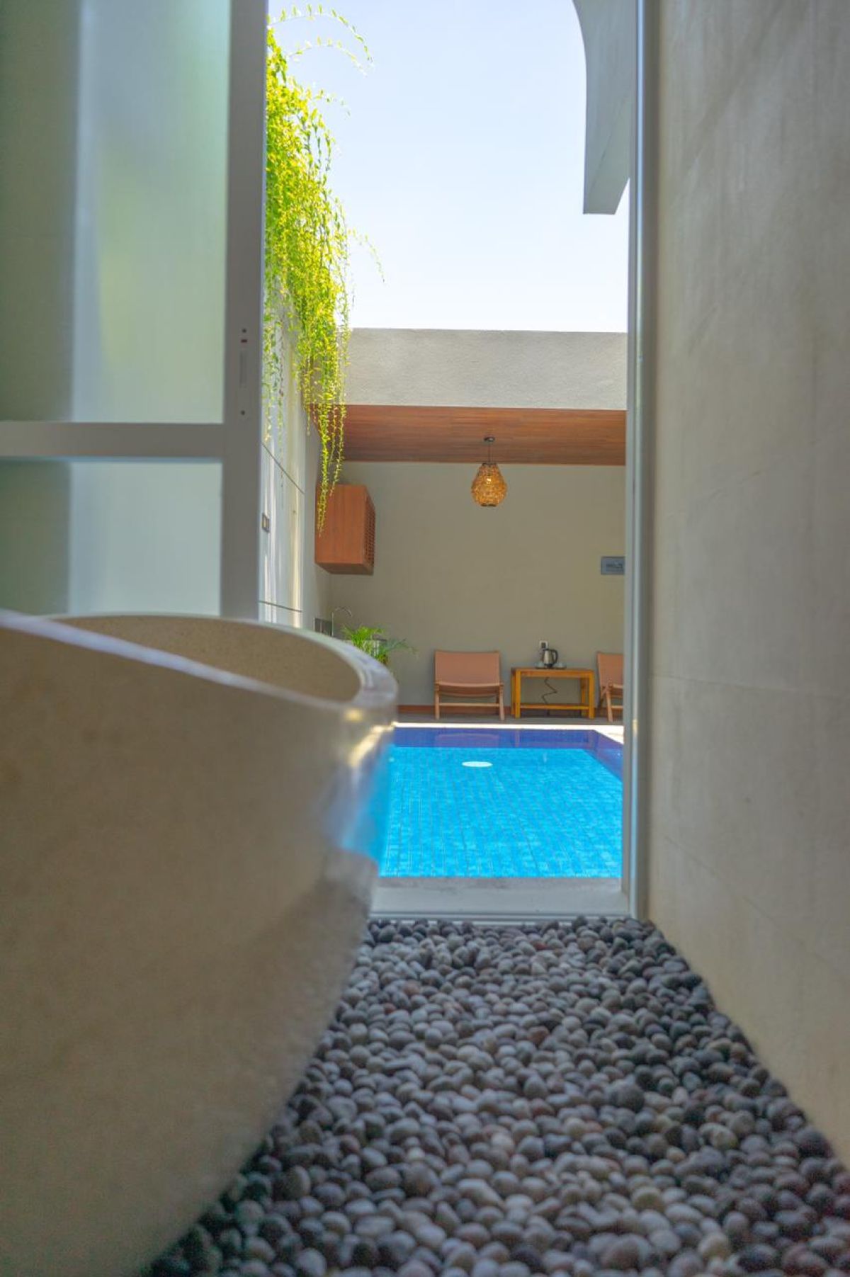 Modern interior with white bathtub and open archway leading to a private blue infinity pool, wooden lounge chairs, hanging lantern, and lush green vines in a serene courtyard. - Bali Villas