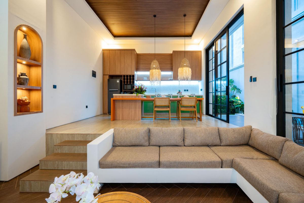 Modern tropical open-plan living room and kitchen with white L-shaped sofa, rattan accents, pendant lights, wooden island bar, and lush greenery views. - Bali Villas