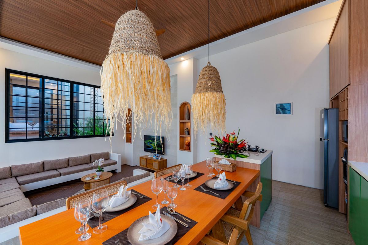 Modern tropical open-plan living room and kitchen with white L-shaped sofa, rattan accents, pendant lights, wooden island bar, and lush greenery views. - Bali Villas