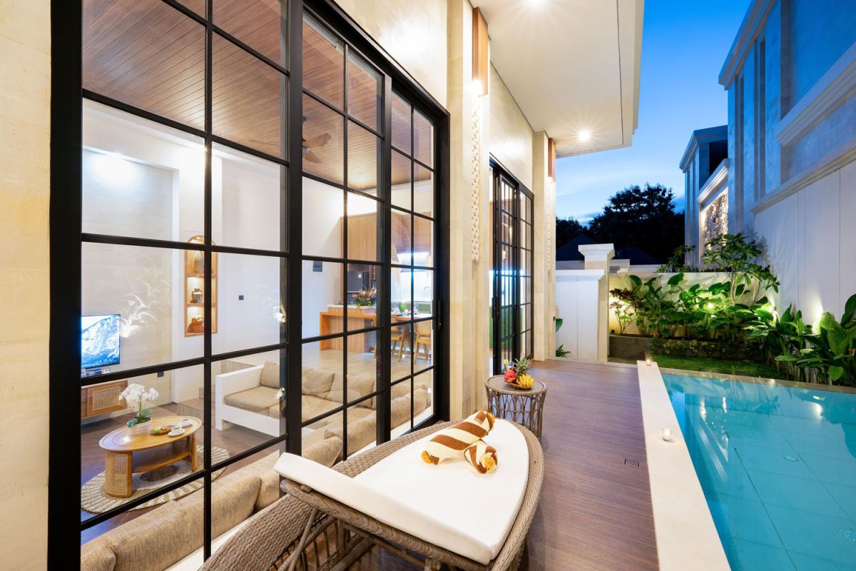 Modern tropical open-plan living room and kitchen with white L-shaped sofa, rattan accents, pendant lights, wooden island bar, and lush greenery views. - Bali Villas