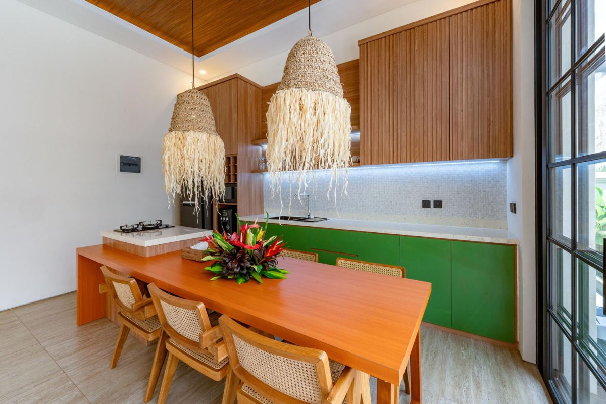Modern tropical open-plan living room and kitchen with white L-shaped sofa, rattan accents, pendant lights, wooden island bar, and lush greenery views. - Bali Villas
