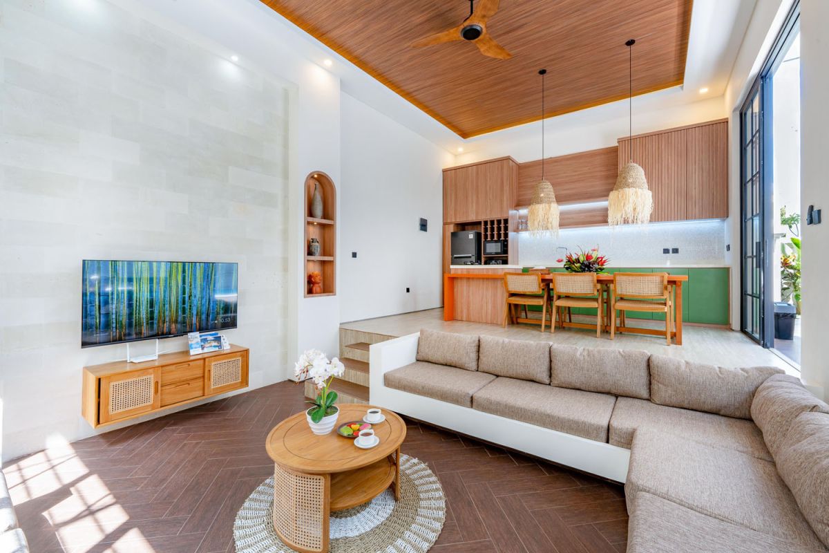 Modern tropical open-plan living room and kitchen with white L-shaped sofa, rattan accents, pendant lights, wooden island bar, and lush greenery views. - Bali Villas
