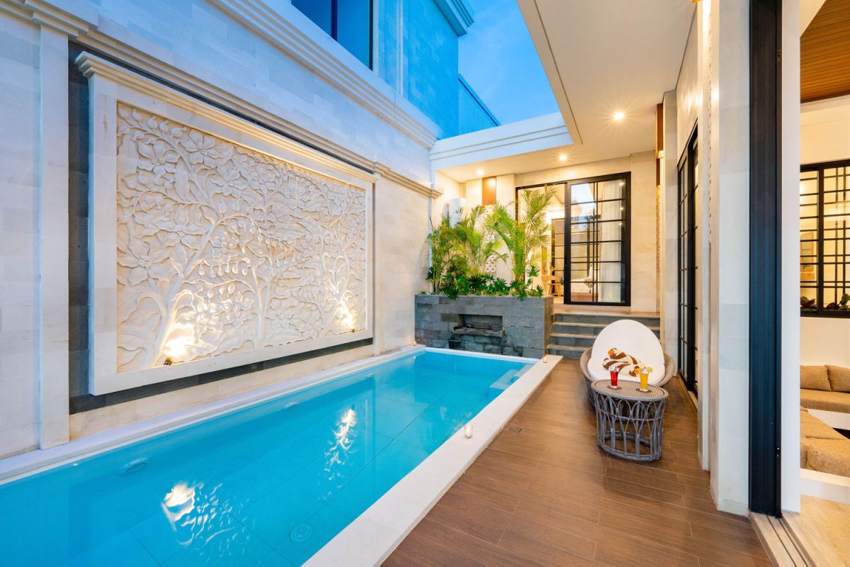 Modern tropical open-plan living room and kitchen with white L-shaped sofa, rattan accents, pendant lights, wooden island bar, and lush greenery views. - Bali Villas