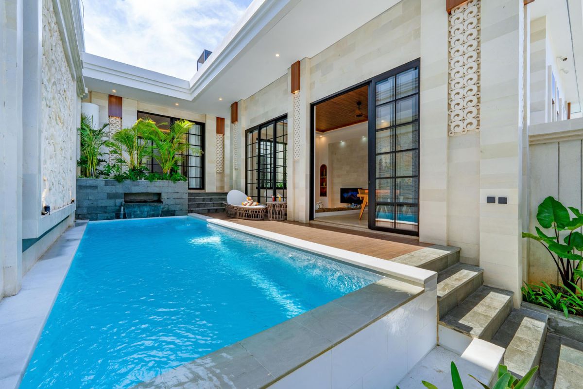 Modern tropical open-plan living room and kitchen with white L-shaped sofa, rattan accents, pendant lights, wooden island bar, and lush greenery views. - Bali Villas
