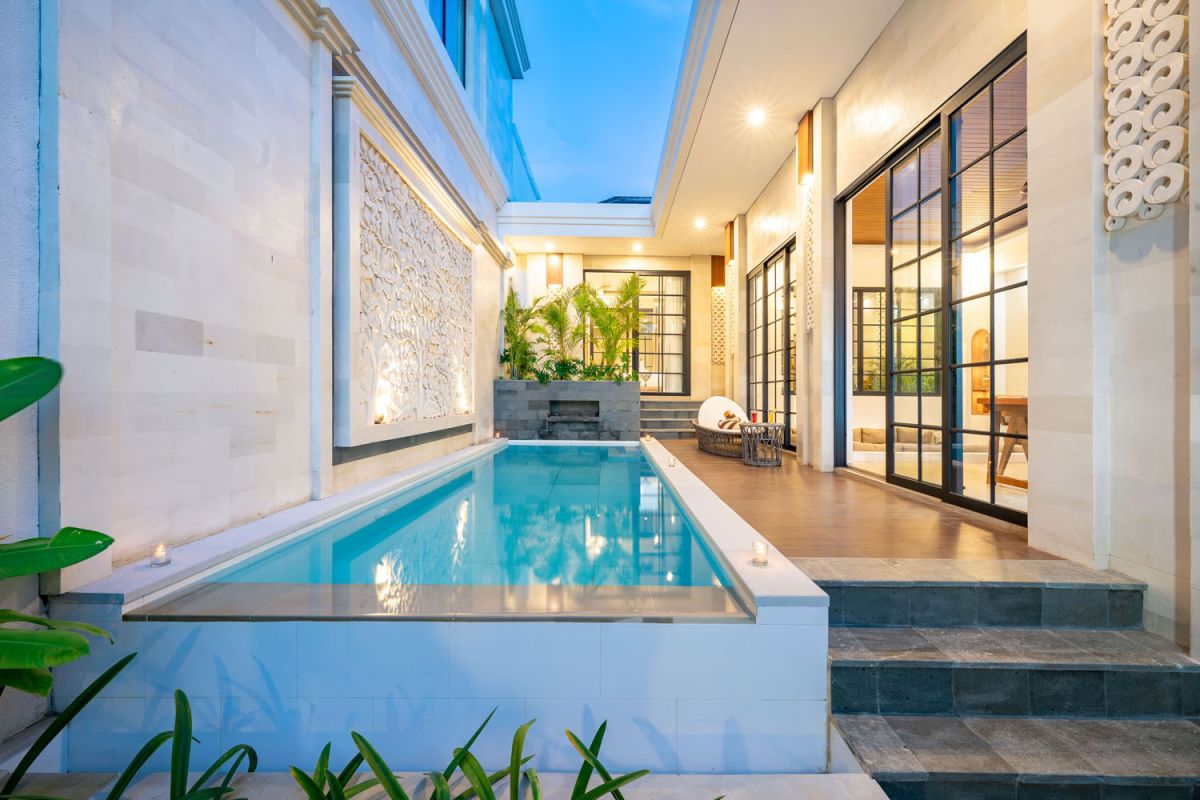 Modern tropical open-plan living room and kitchen with white L-shaped sofa, rattan accents, pendant lights, wooden island bar, and lush greenery views. - Bali Villas