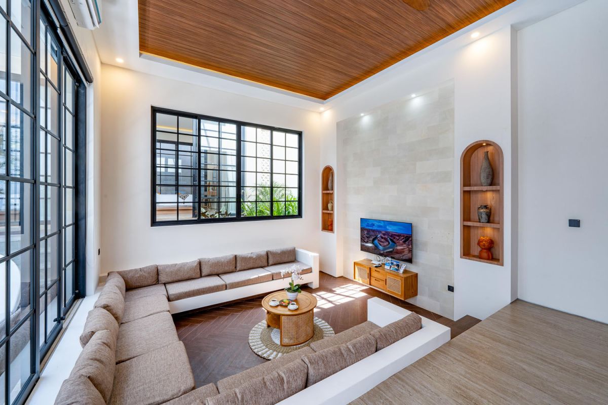 Modern tropical open-plan living room and kitchen with white L-shaped sofa, rattan accents, pendant lights, wooden island bar, and lush greenery views. - Bali Villas