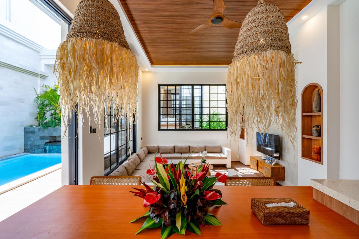 Modern tropical open-plan living room and kitchen with white L-shaped sofa, rattan accents, pendant lights, wooden island bar, and lush greenery views. - Bali Villas