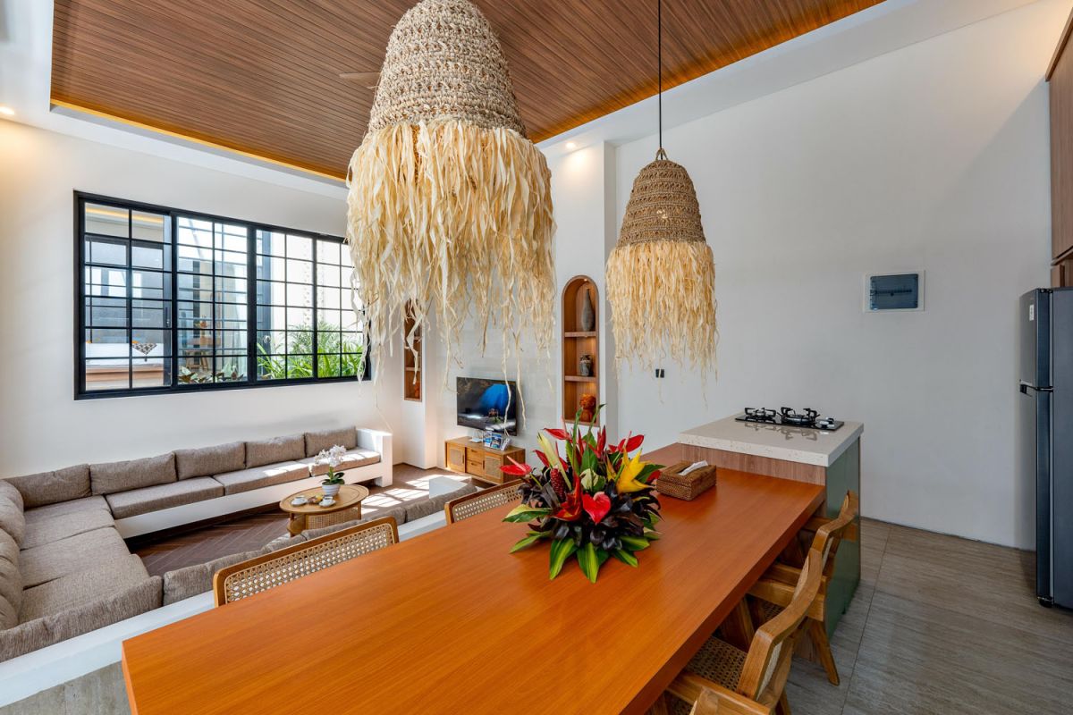 Modern tropical open-plan living room and kitchen with white L-shaped sofa, rattan accents, pendant lights, wooden island bar, and lush greenery views. - Bali Villas