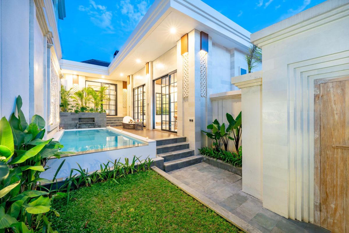 Modern tropical open-plan living room and kitchen with white L-shaped sofa, rattan accents, pendant lights, wooden island bar, and lush greenery views. - Bali Villas