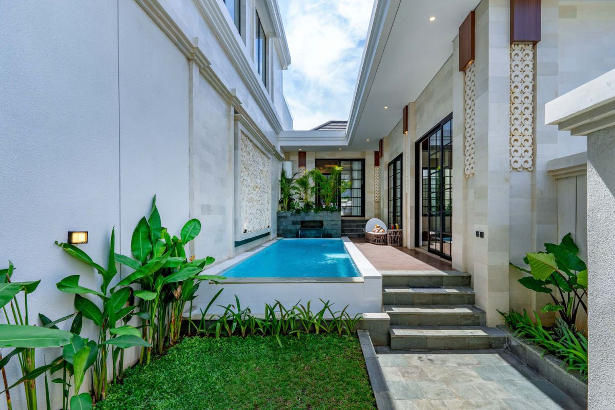 Modern tropical open-plan living room and kitchen with white L-shaped sofa, rattan accents, pendant lights, wooden island bar, and lush greenery views. - Bali Villas
