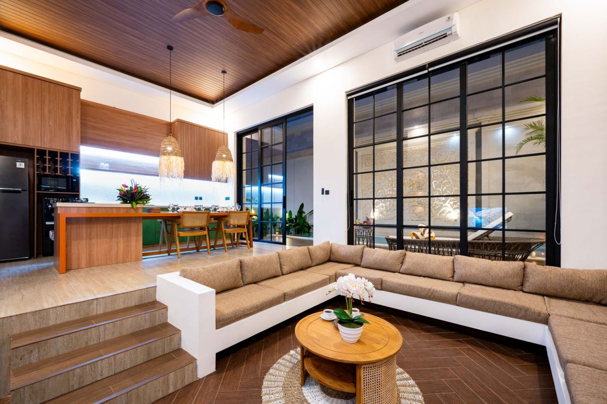 Modern tropical open-plan living room and kitchen with white L-shaped sofa, rattan accents, pendant lights, wooden island bar, and lush greenery views. - Bali Villas