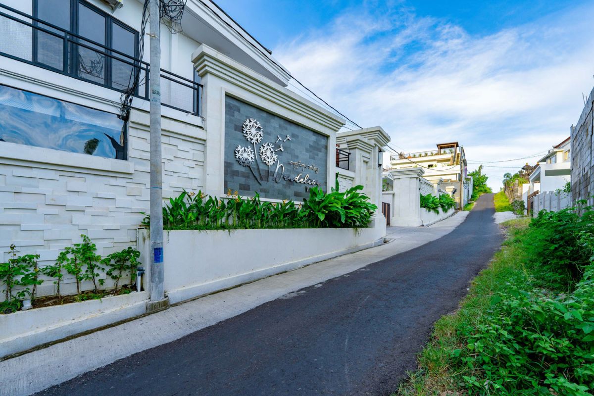 Modern tropical open-plan living room and kitchen with white L-shaped sofa, rattan accents, pendant lights, wooden island bar, and lush greenery views. - Bali Villas
