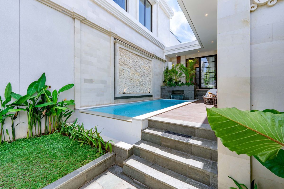 Modern tropical open-plan living room and kitchen with white L-shaped sofa, rattan accents, pendant lights, wooden island bar, and lush greenery views. - Bali Villas