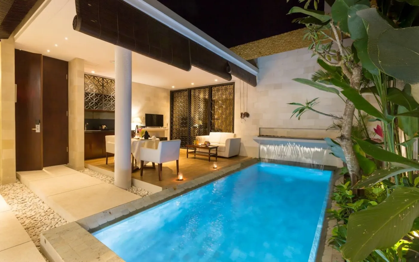 Luxurious Bali villa courtyard at night: illuminated infinity pool, outdoor dining table, sofa seating, and lush tropical plants under a wooden pavilion. - Bali Villas