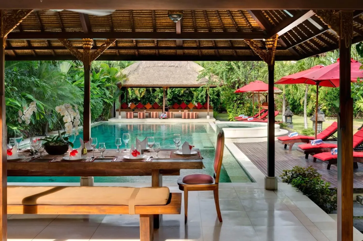 Luxurious open-air Bali pavilion with poolside wooden dining table, red loungers, and tropical garden. - Bali Villas