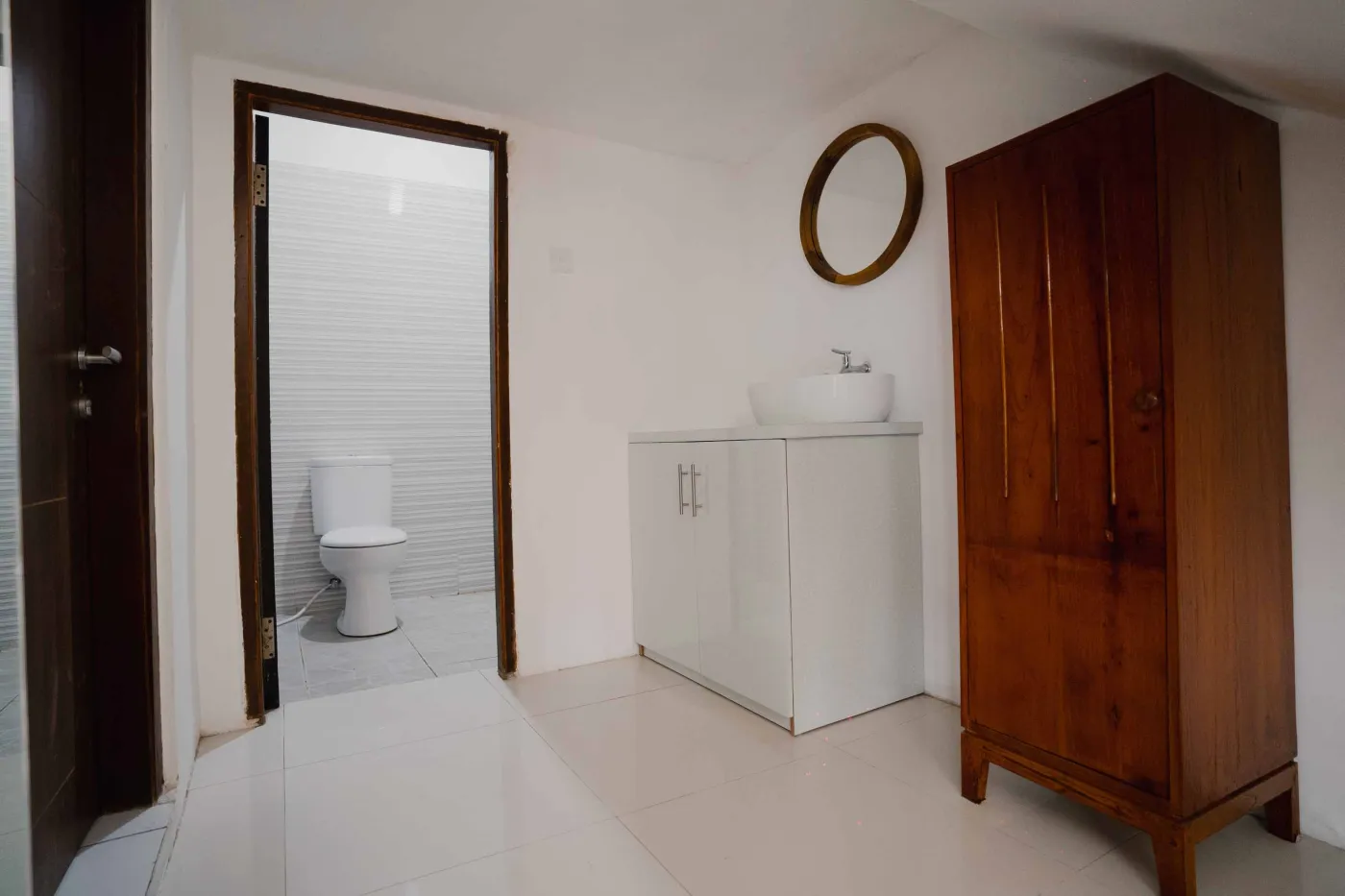 Modern bathroom with white toilet, sink vanity, wooden wardrobe, and round mirror on white brick walls. - Bali Villas