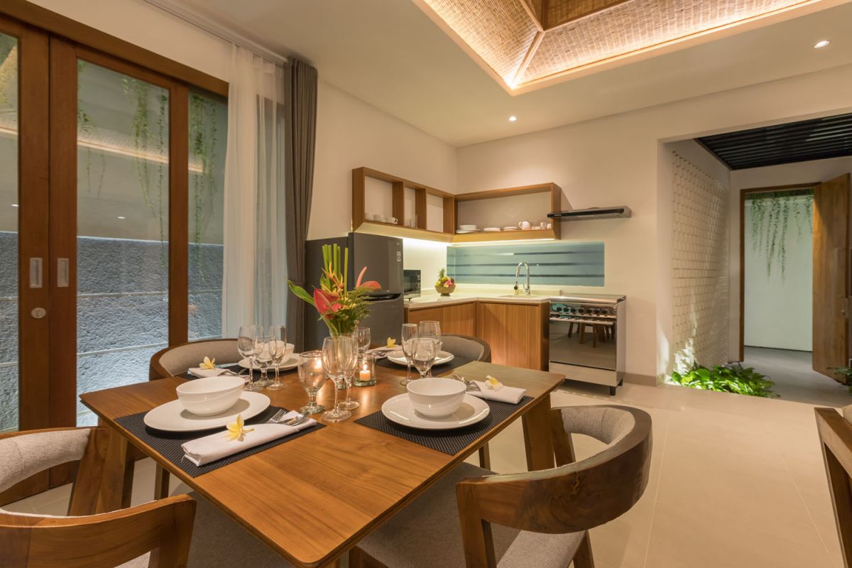 Modern wooden kitchen and dining area with set table, tropical flowers, and sliding glass doors to a lush balcony. - Bali Villas