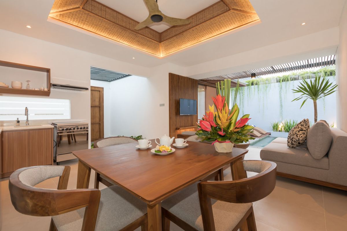 Modern wooden kitchen and dining area with set table, tropical flowers, and sliding glass doors to a lush balcony. - Bali Villas