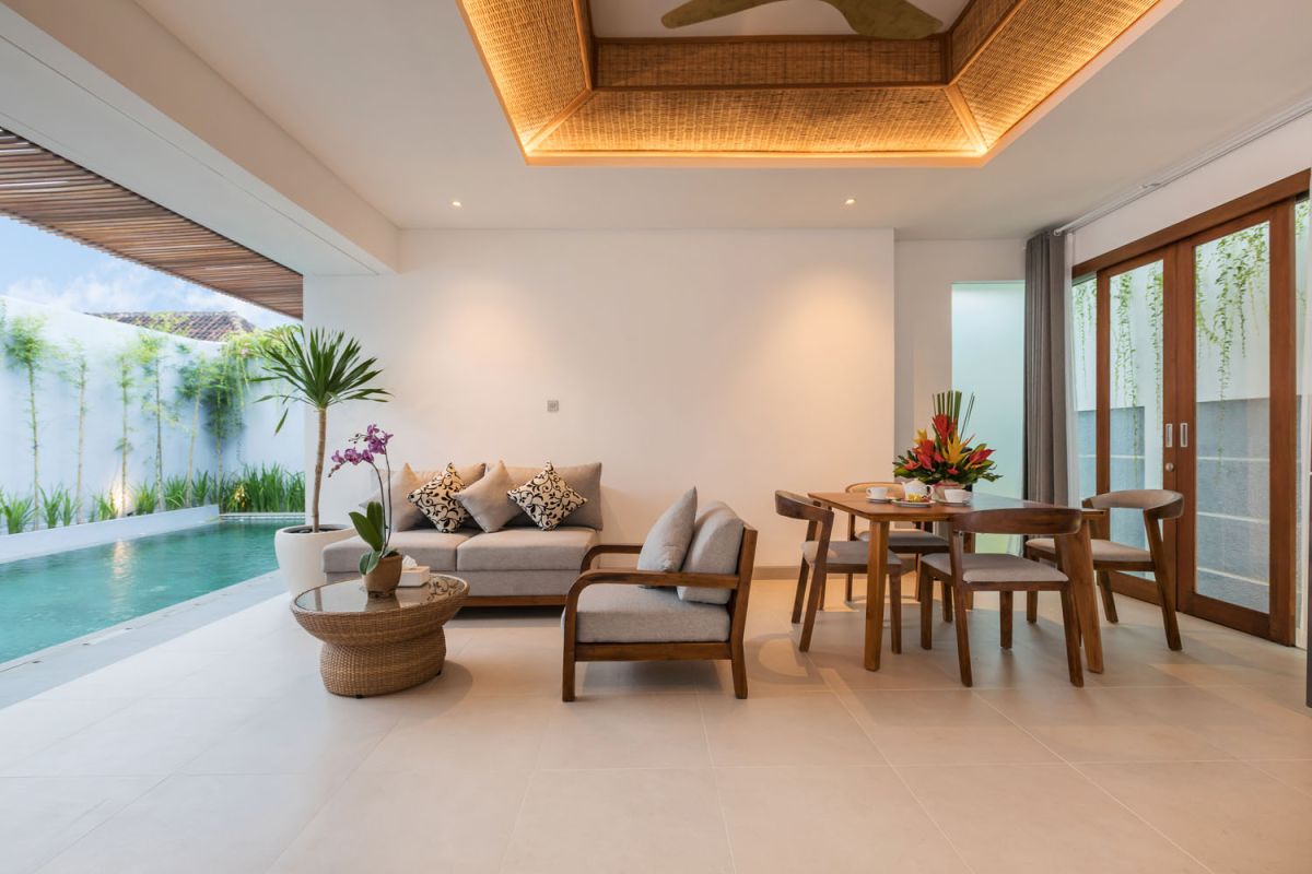 Modern wooden kitchen and dining area with set table, tropical flowers, and sliding glass doors to a lush balcony. - Bali Villas