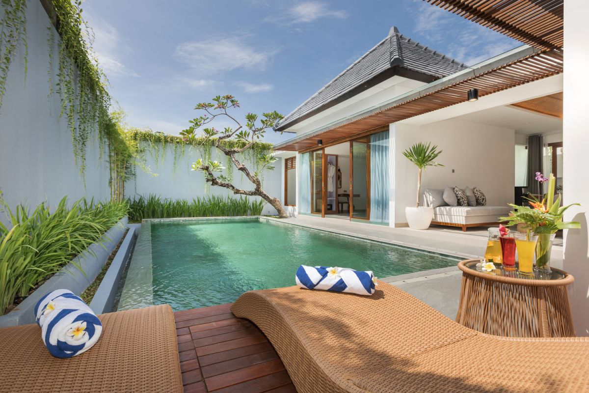 Luxury Balinese villa poolside with turquoise infinity pool, rattan lounge chairs, tropical plants, and cocktails under a clear sky. - Bali Villas