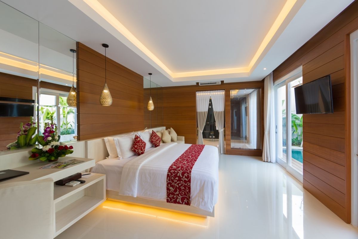 Spacious Balinese villa bedroom with wooden walls, white king bed, red accents, and sliding doors to a private infinity pool. - Bali Villas