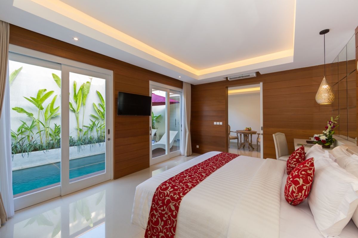 Spacious Balinese villa bedroom with wooden walls, white king bed, red accents, and sliding doors to a private infinity pool. - Bali Villas