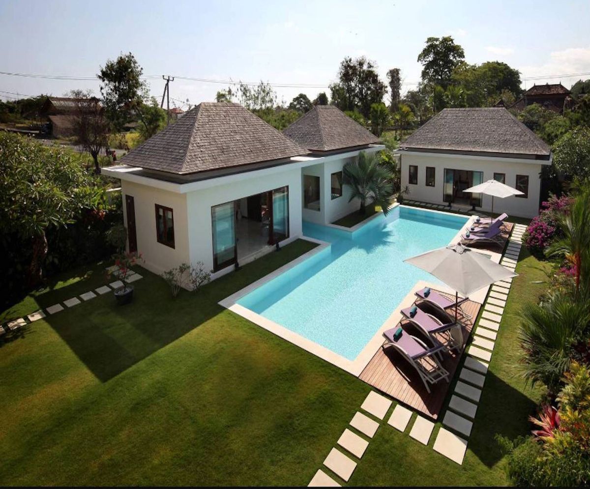 Modern tropical villa with infinity pool, purple loungers, and lush gardens under a clear blue sky. - Bali Villas
