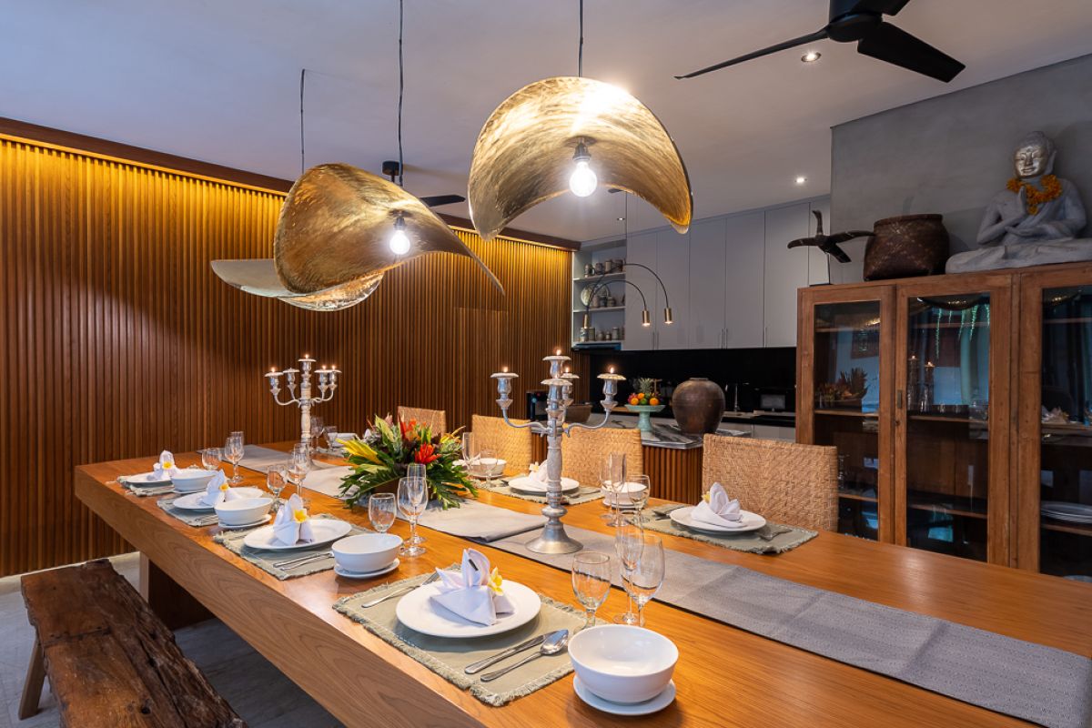 Elegant Balinese dining room with a set wooden table, rattan chairs, tropical floral centerpiece, hanging leaf lamps, and open doors to an outdoor pool. - Bali Villas