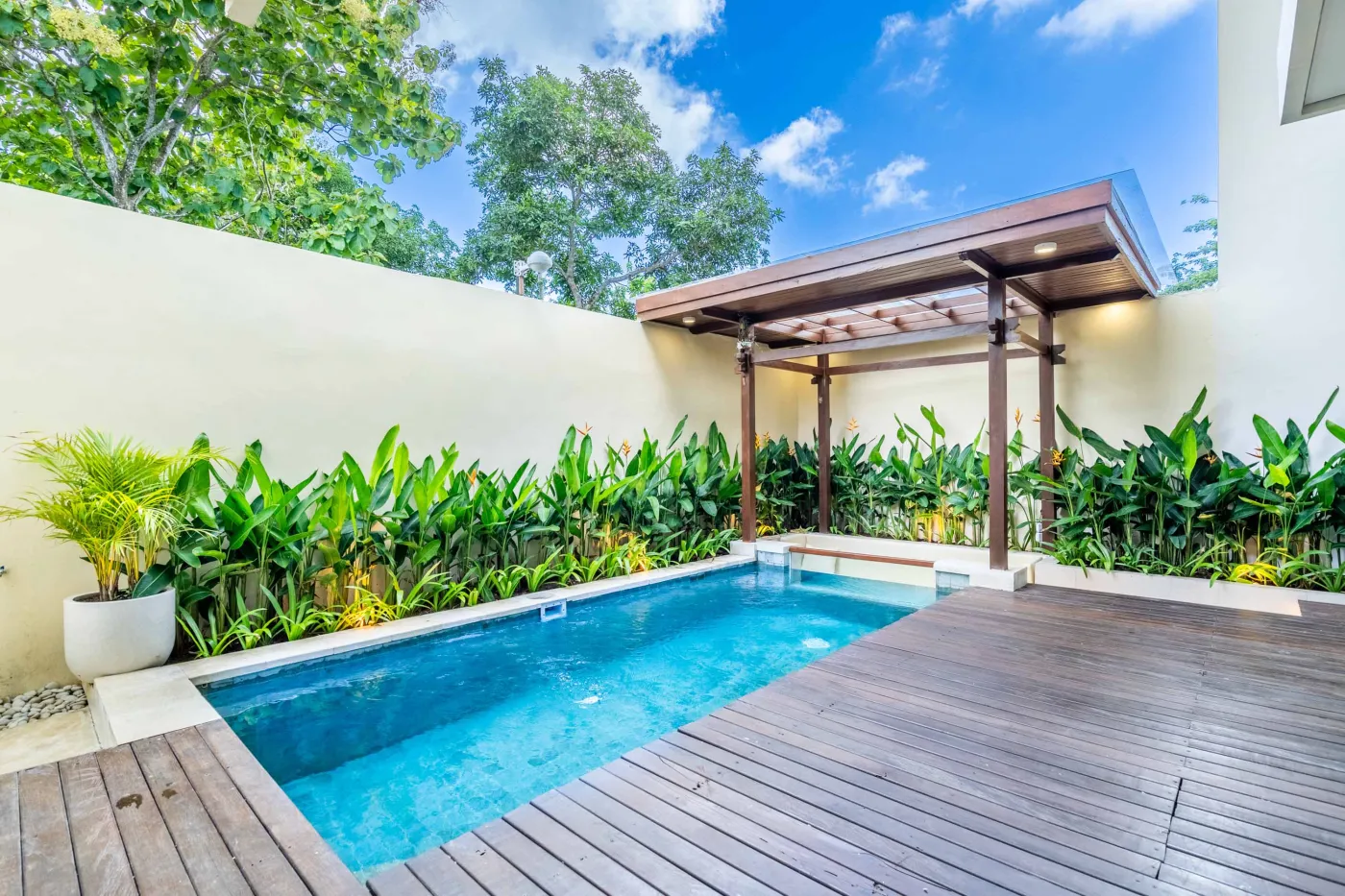 Modern minimalist bathroom featuring a freestanding white bathtub, sleek vanity sink with black faucet, round wooden mirror, and tropical plant-filled window. - Bali Villas