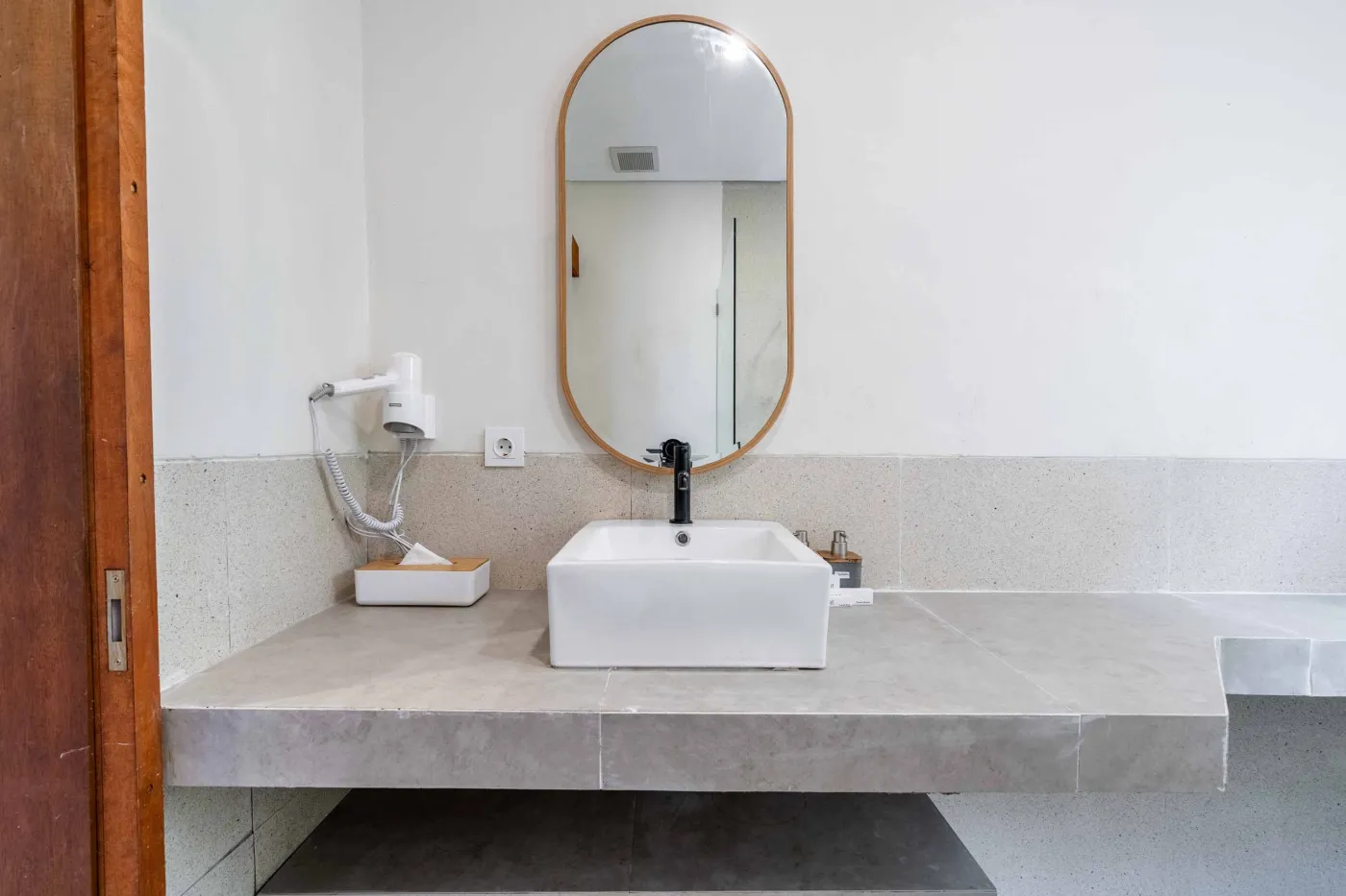 Modern minimalist bathroom featuring a freestanding white bathtub, sleek vanity sink with black faucet, round wooden mirror, and tropical plant-filled window. - Bali Villas