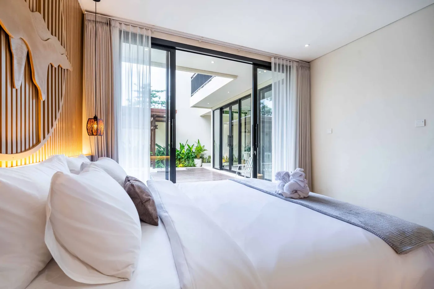Modern minimalist bathroom featuring a freestanding white bathtub, sleek vanity sink with black faucet, round wooden mirror, and tropical plant-filled window. - Bali Villas